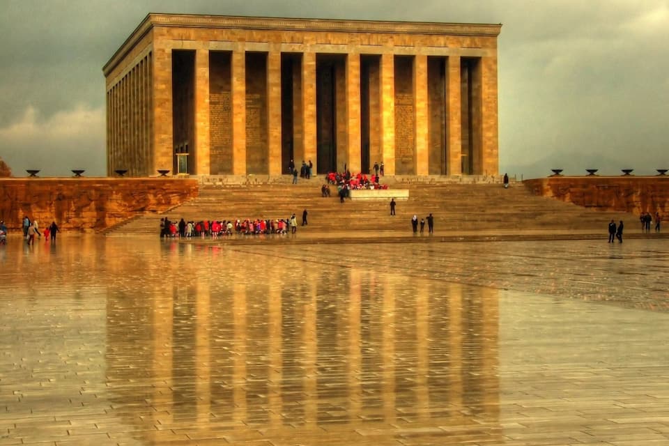 Un edifici gran i rectangular amb columnes altes, una ampla piscina reflectant i un grup de persones reunides a les escales.