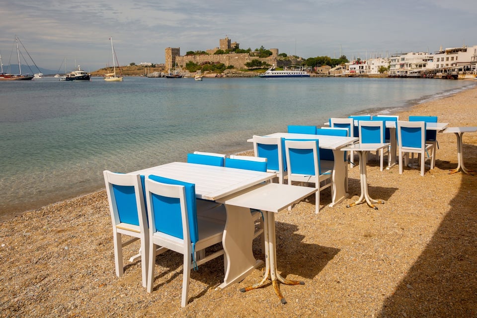En strand med vita bord och blå stolar, ett slott i bakgrunden och båtar på vattnet.