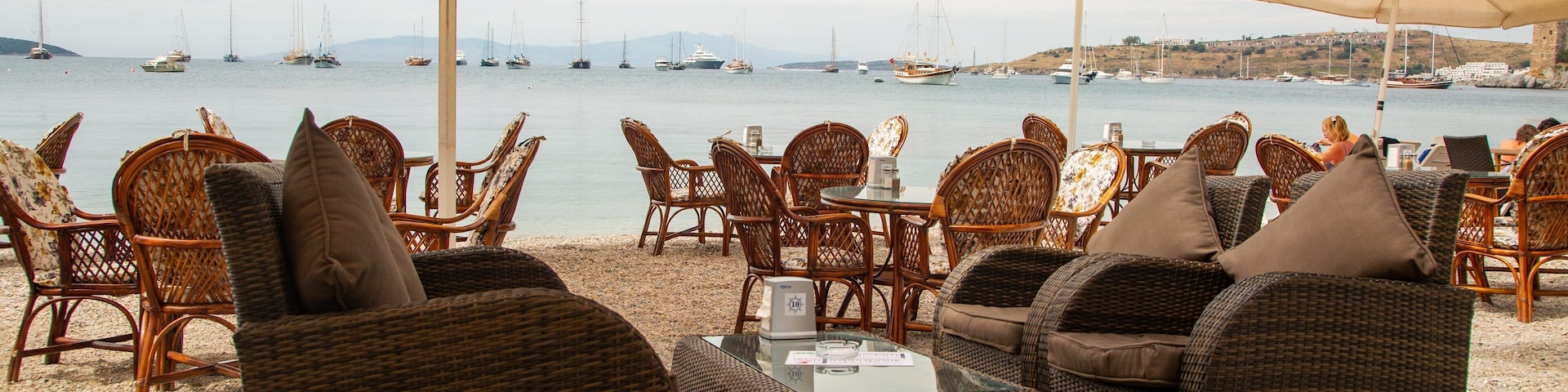 Sillas y mesas de mimbre en una playa con sombrillas, con vista a un puerto deportivo con barcos.
