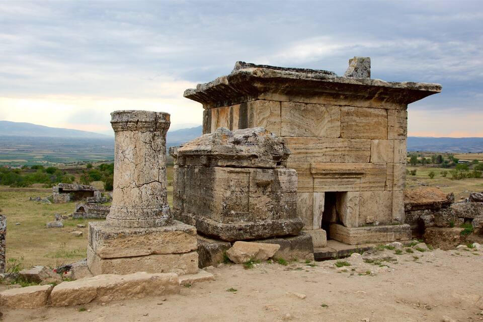 Ancient stone ruins with columns and a small building, surrounded by a rural landscape with mountains in the background.