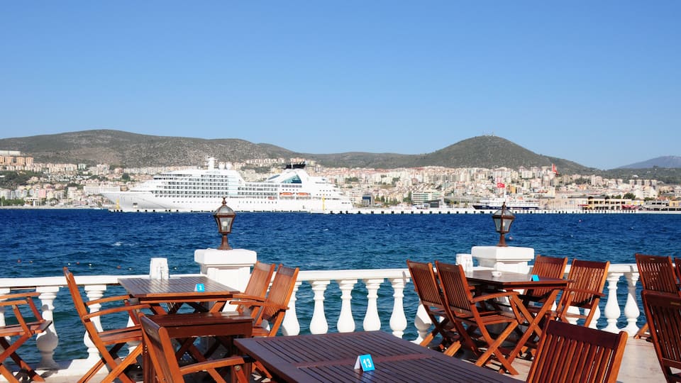 A seaside café with wooden tables and chairs, overlooking a large cruise ship and a cityscape with hills in the background.