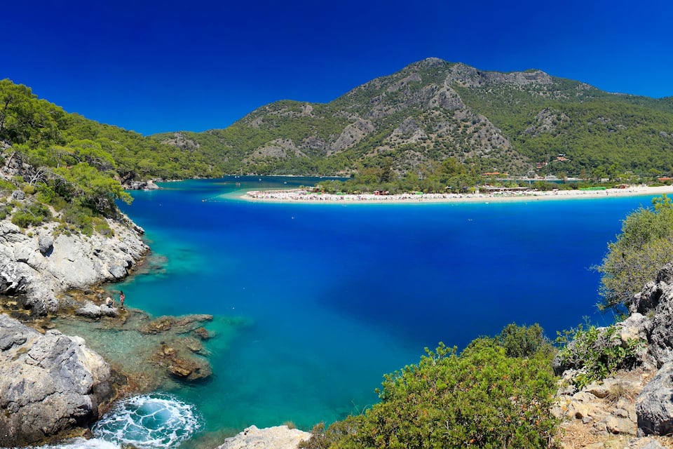 A coastal landscape with clear turquoise waters, rocky cliffs, and a mountainous backdrop.