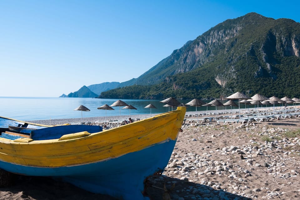 A beach with a yellow and blue boat, beach umbrellas, and a mountain in the background.