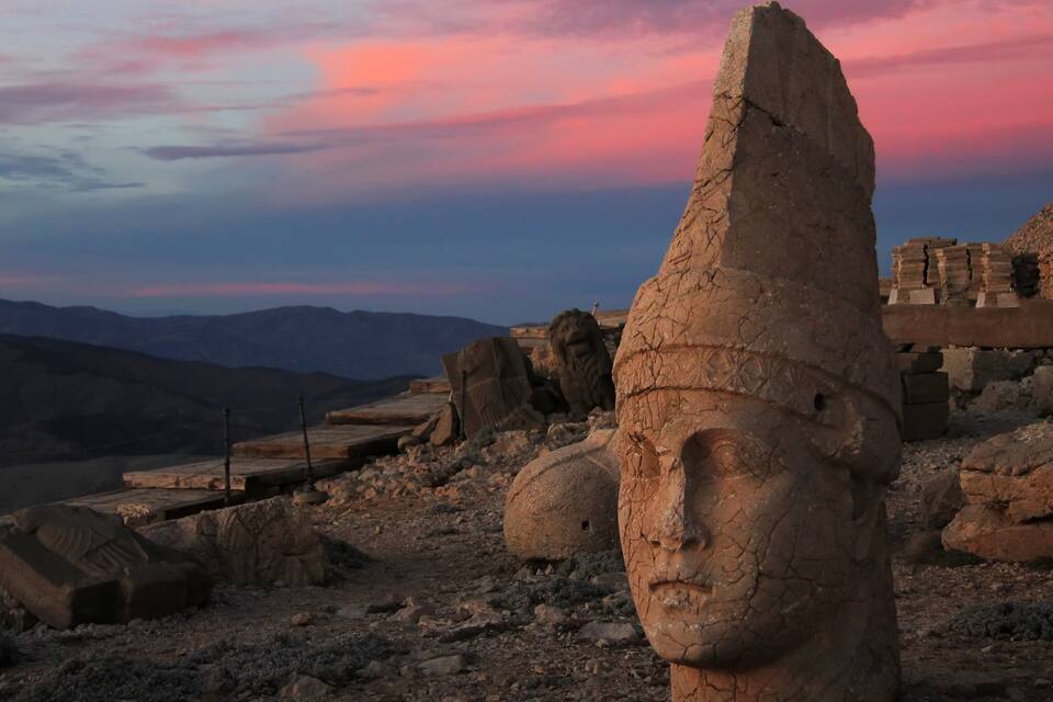 A large stone head sculpture in a desert landscape with mountains in the background and a colorful sky.