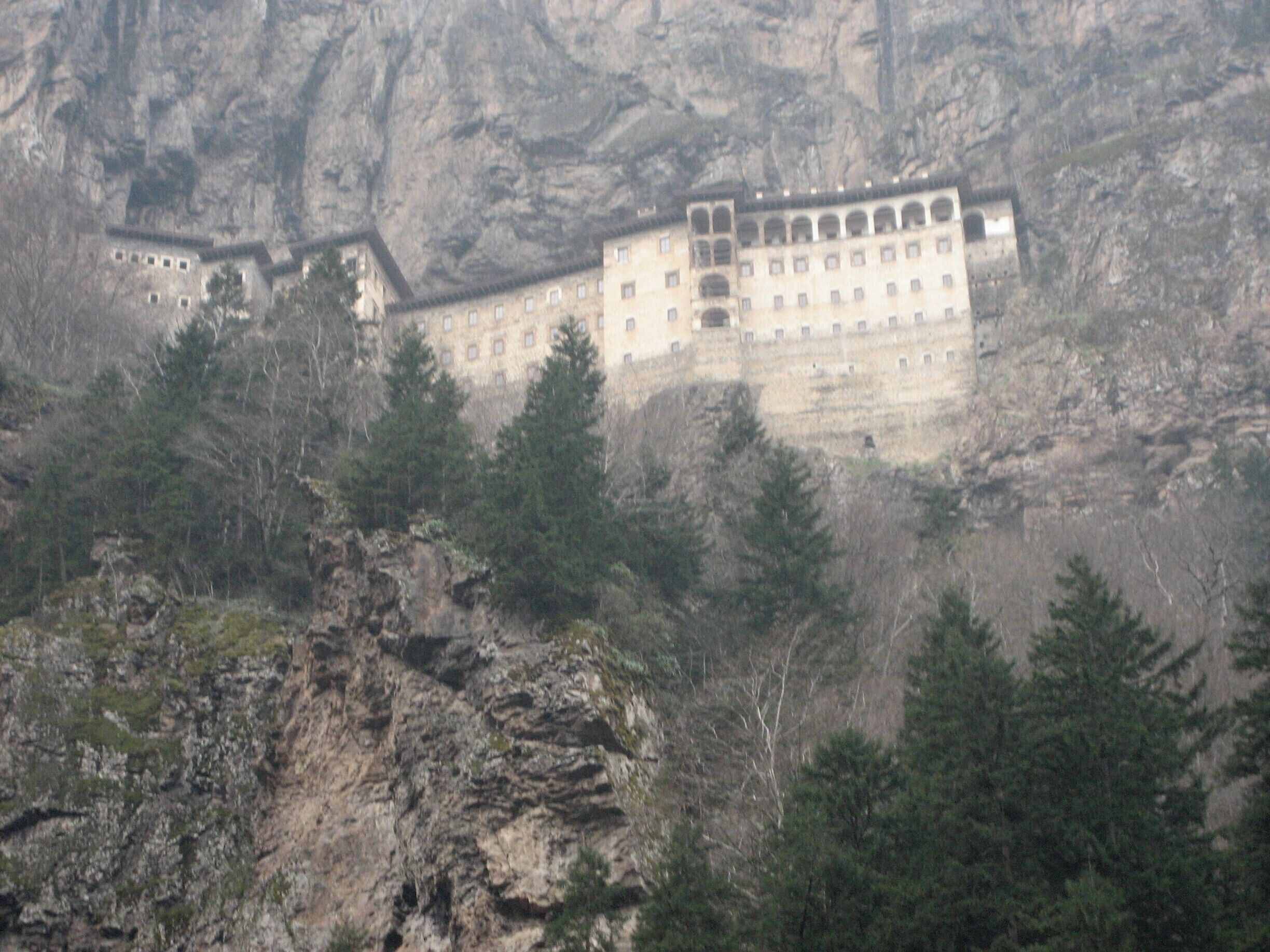 A large, multi-story building with a flat roof, surrounded by rocky cliffs and dense forest.