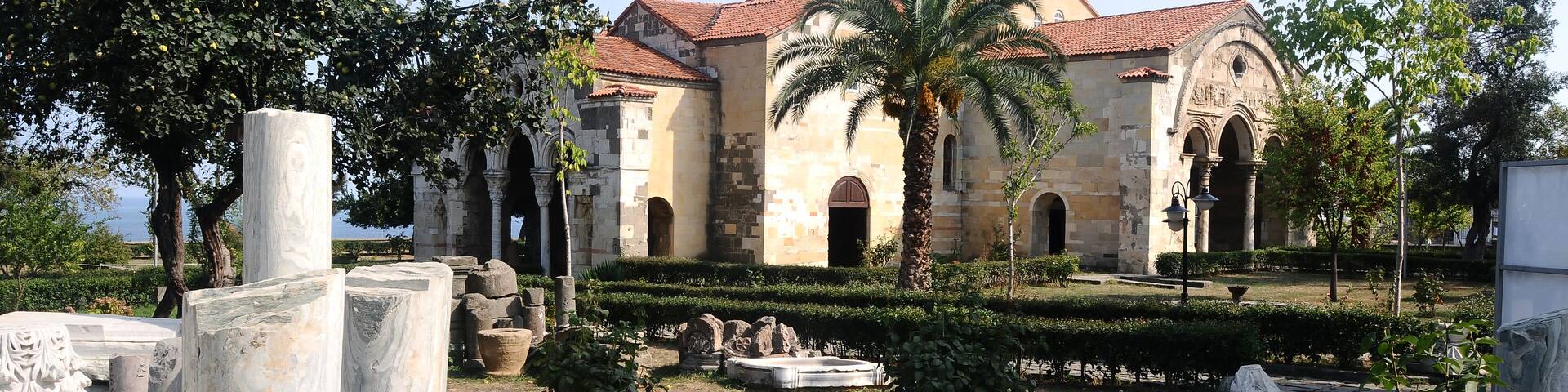 A historic building with a dome and arched windows, surrounded by a well-maintained garden with palm trees and stone columns.