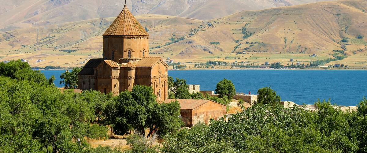 A historic church with a bell tower, surrounded by lush greenery and a lake, with a mountain in the background.