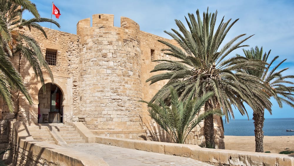Une forteresse historique en pierre, avec un drapeau, des palmiers et une plage en arrière-plan.