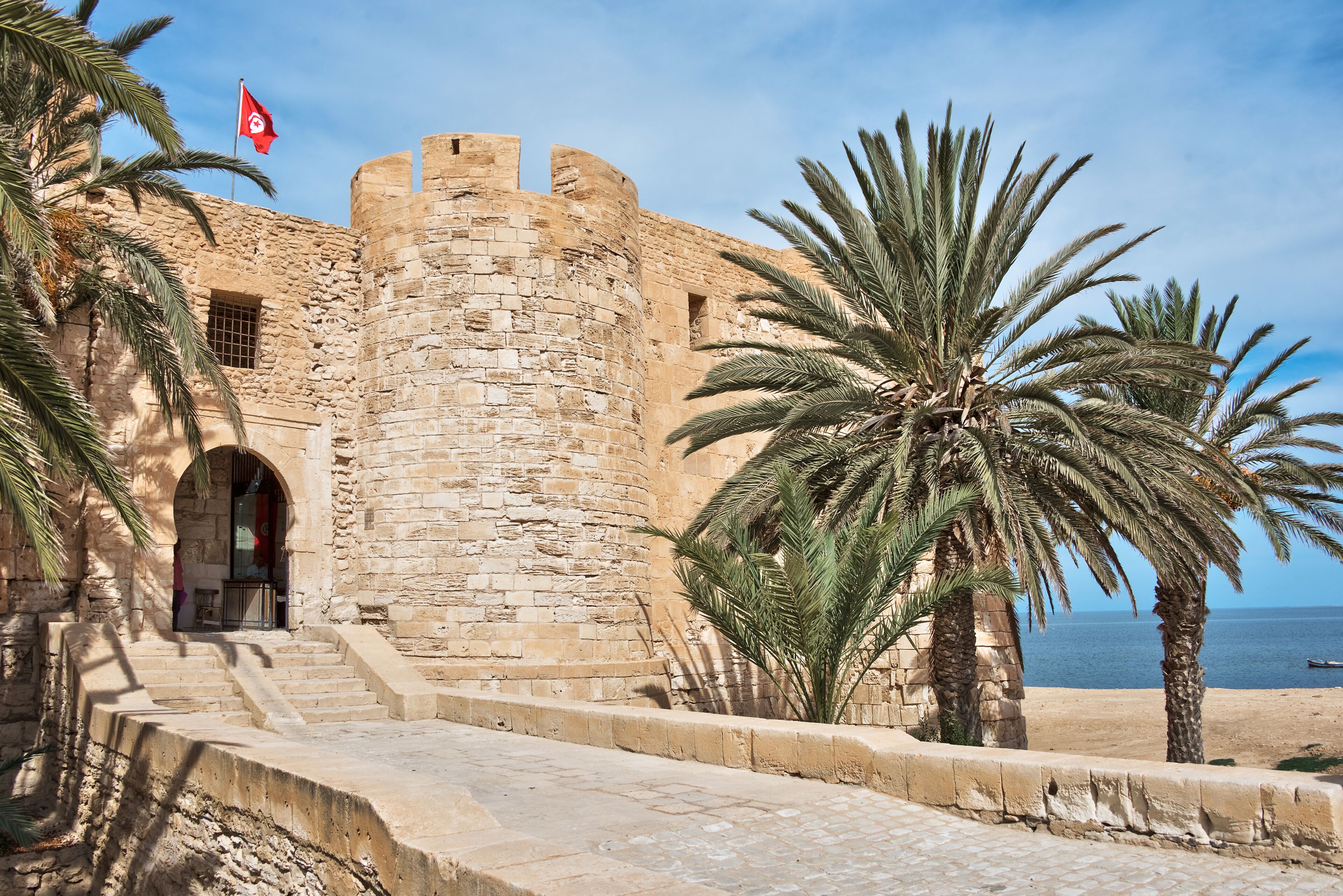 A historic stone fortress with a flag, palm trees, and a beach in the background.