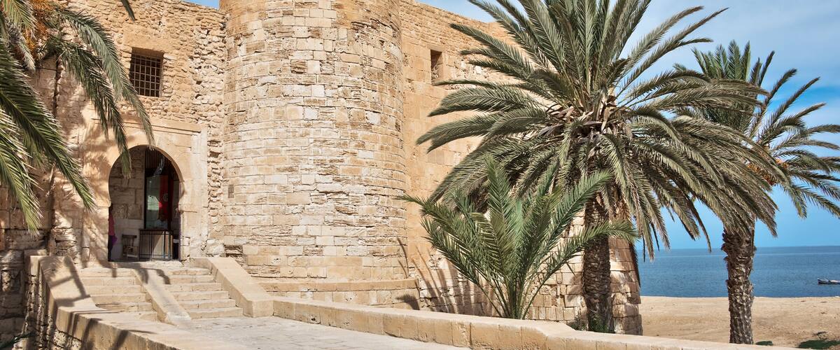 A historic stone fortress with a flag, palm trees, and a beach in the background.