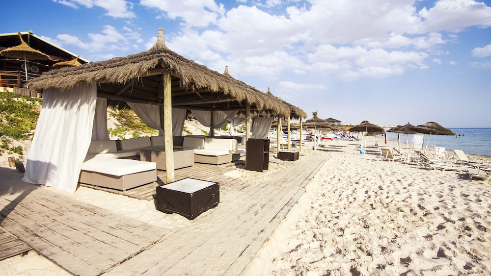 A beach with thatched-roof huts, wooden deck, and lounge chairs.