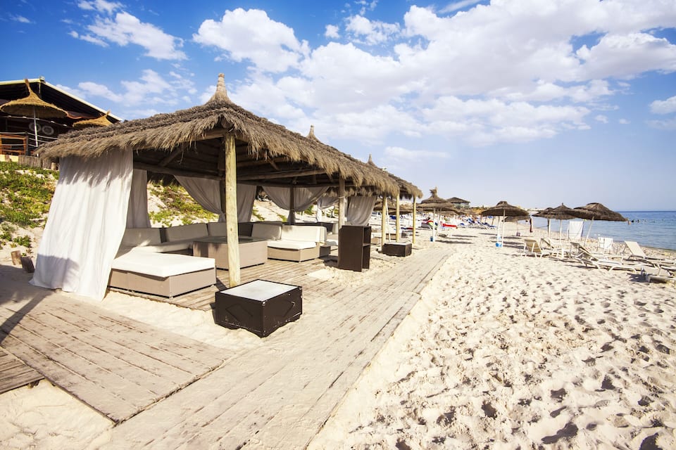 A beach with thatched-roof huts, wooden deck, and lounge chairs.