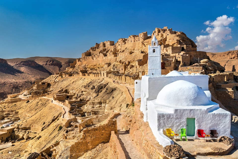 A white mosque with a minaret, set against a mountainous backdrop.