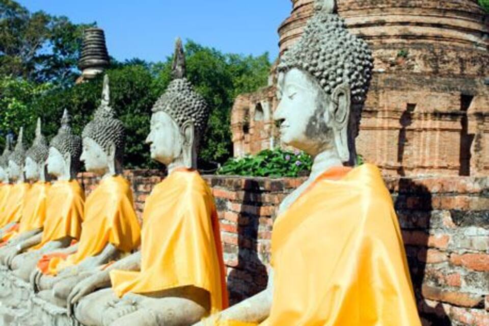 A row of seated Buddha statues with yellow cloths, set against a brick wall and a large stupa in the background.