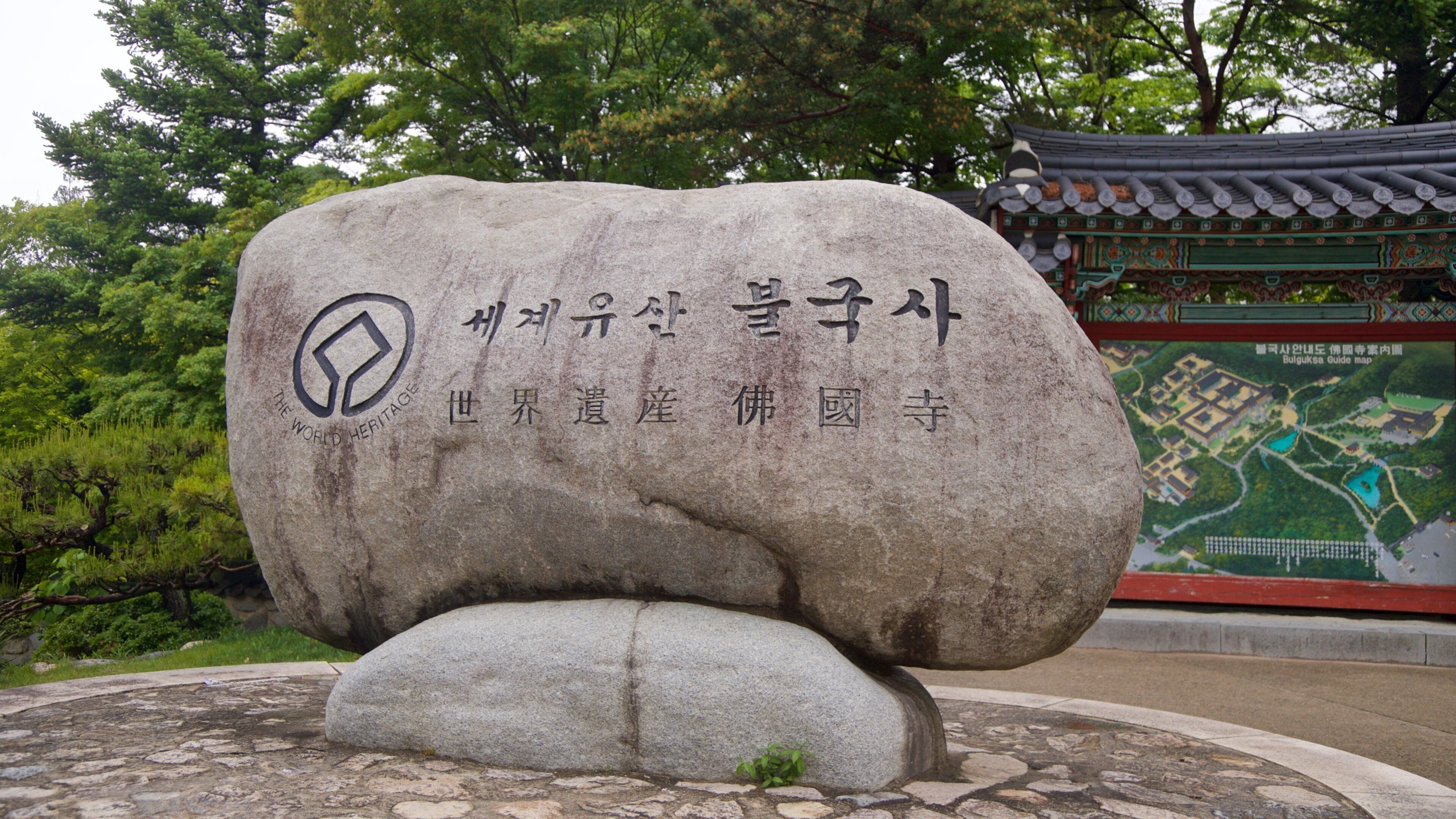 A large stone monument with inscriptions, surrounded by greenery and a traditional building in the background.