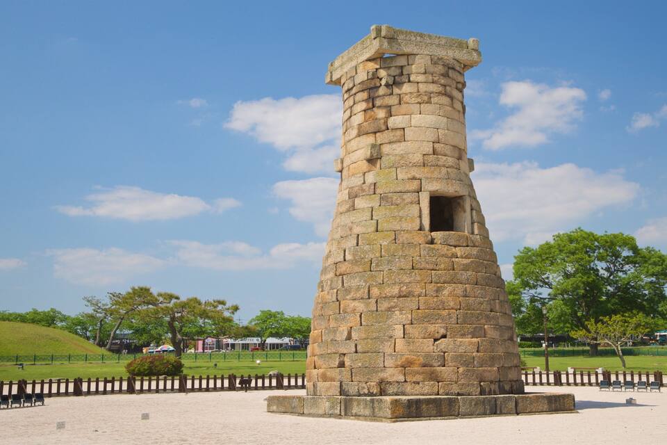 Una torre de piedra con una pequeña abertura en la parte superior, rodeada de una zona arenosa con bancos y árboles.