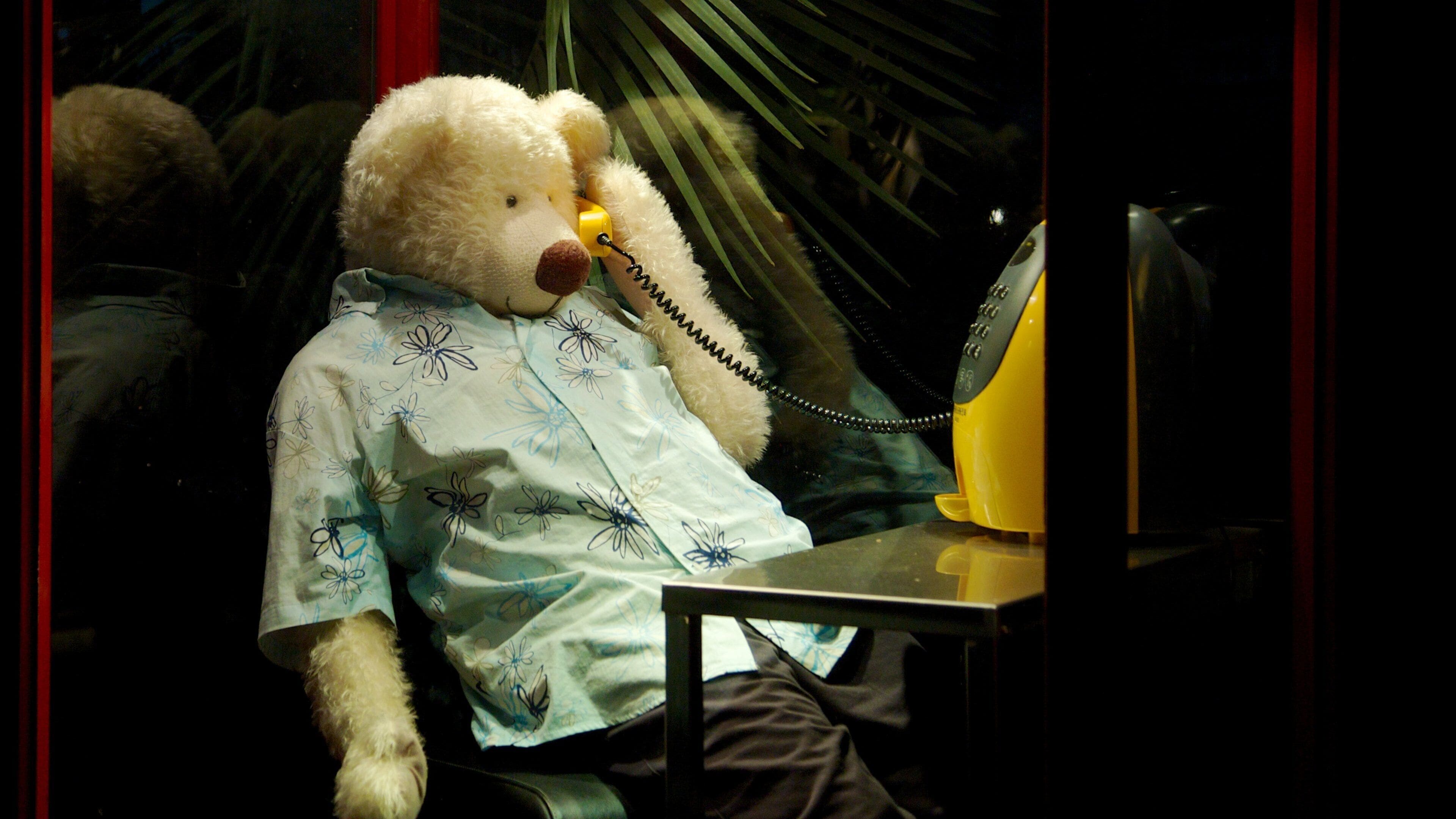 Een teddybeer met een bloemenoverhemd zit aan een tafel met een gele telefoon.