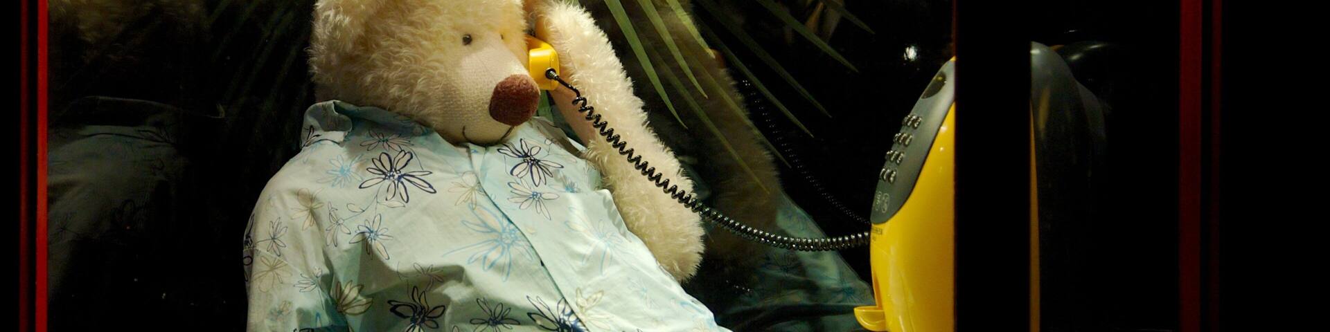 A teddy bear wearing a floral shirt sitting at a table with a yellow telephone.