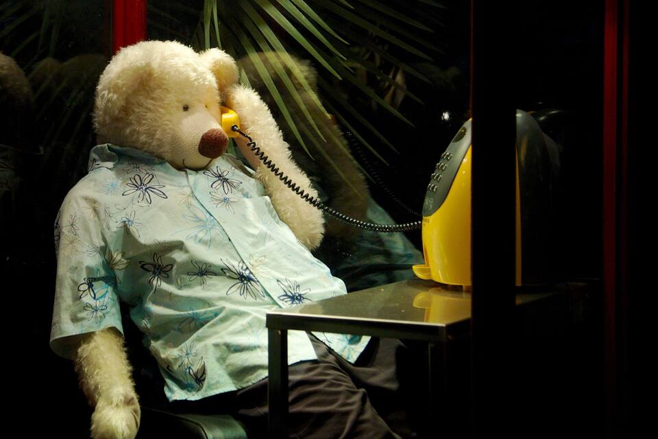Un oso de peluche con una camisa estampada floral sentado a una mesa con un teléfono amarillo.