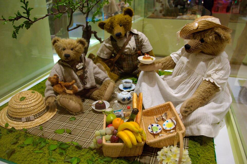 A display of teddy bears dressed in vintage clothing, sitting at a table with tea, biscuits, and fruit.