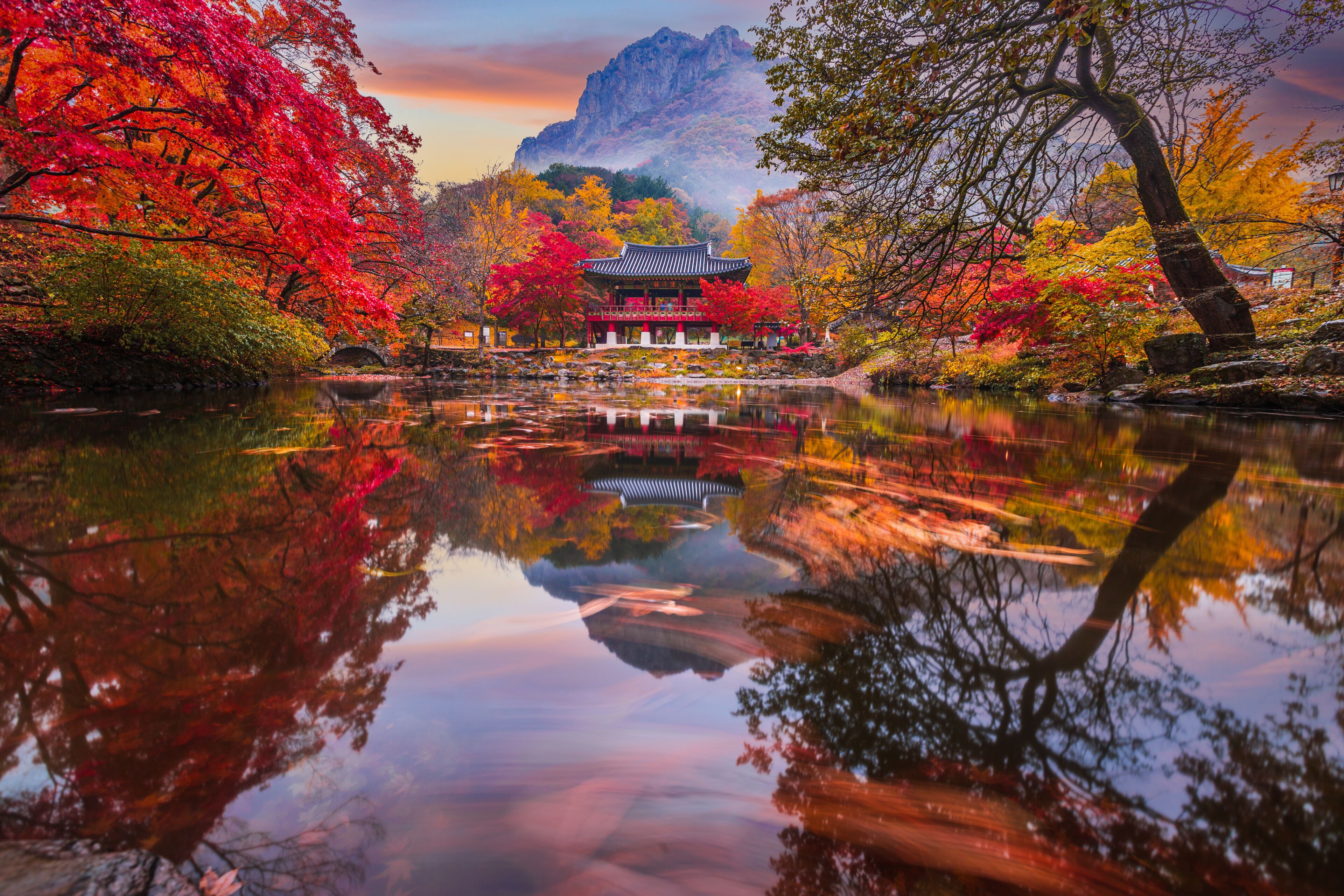 A traditional pavilion by a reflective pond with vibrant autumn foliage and a mountain backdrop.