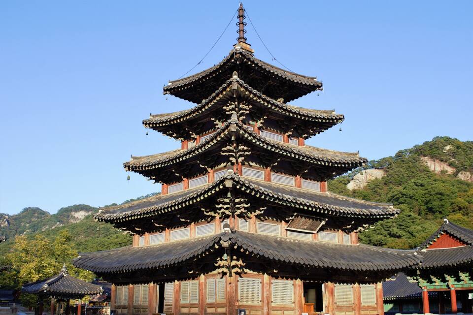 A traditional multi-tiered pagoda with ornate wooden details, set against a backdrop of lush green hills and clear blue sky.