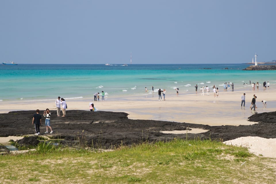 A beach with people walking and standing, a lighthouse in the distance, and a ship on the water.