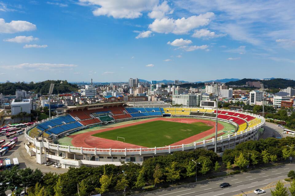 Um estádio grande com gramado verde e pista vermelha, cercado por árvores e edifícios urbanos.
