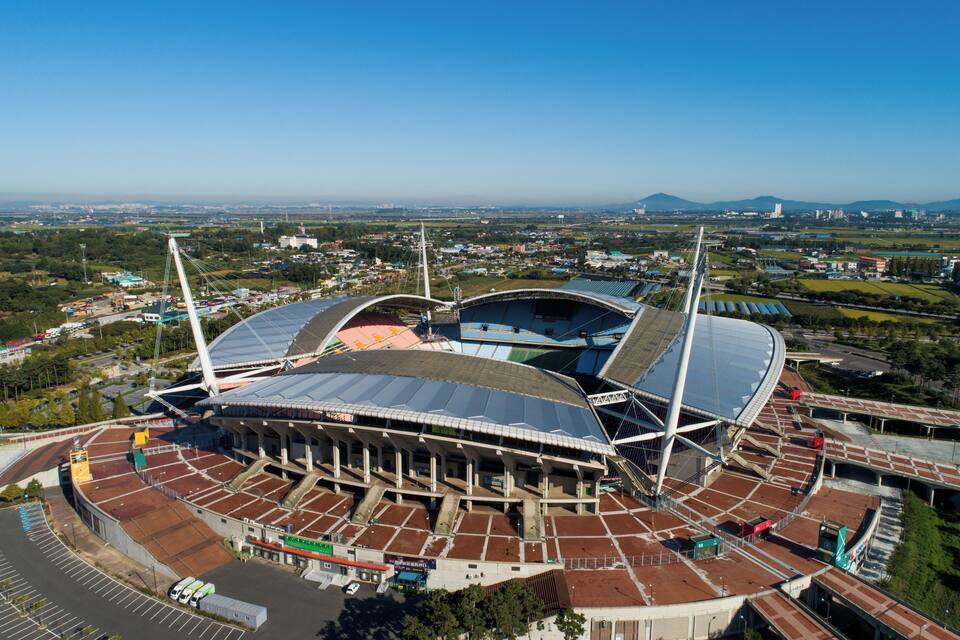 Um estádio grande com uma estrutura de telhado marcante, rodeado por um horizonte urbano e áreas verdes.