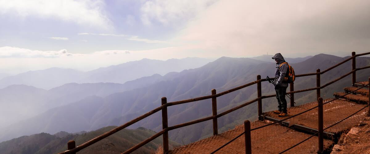 A hiker on a mountain trail with a wooden fence and a view of distant mountains under a cloudy sky.