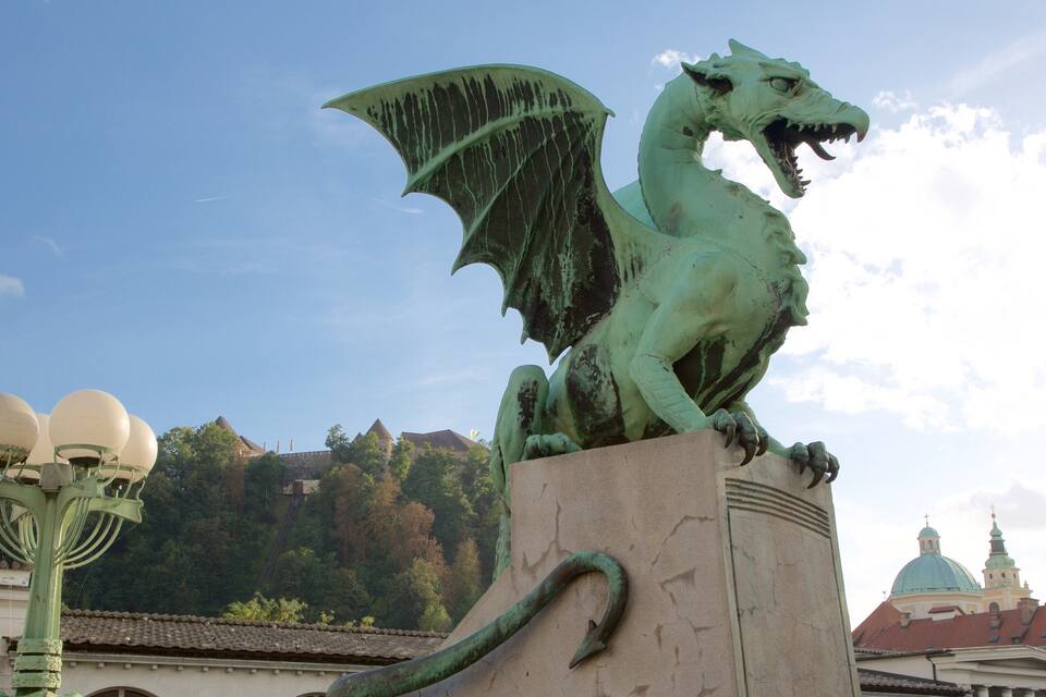 A green dragon statue on a pedestal with a street lamp nearby, set against a backdrop of buildings and a hill with trees.