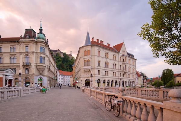 A cobblestone street lined with historic buildings, a bridge with ornate balustrades, and a distant castle on a hill.