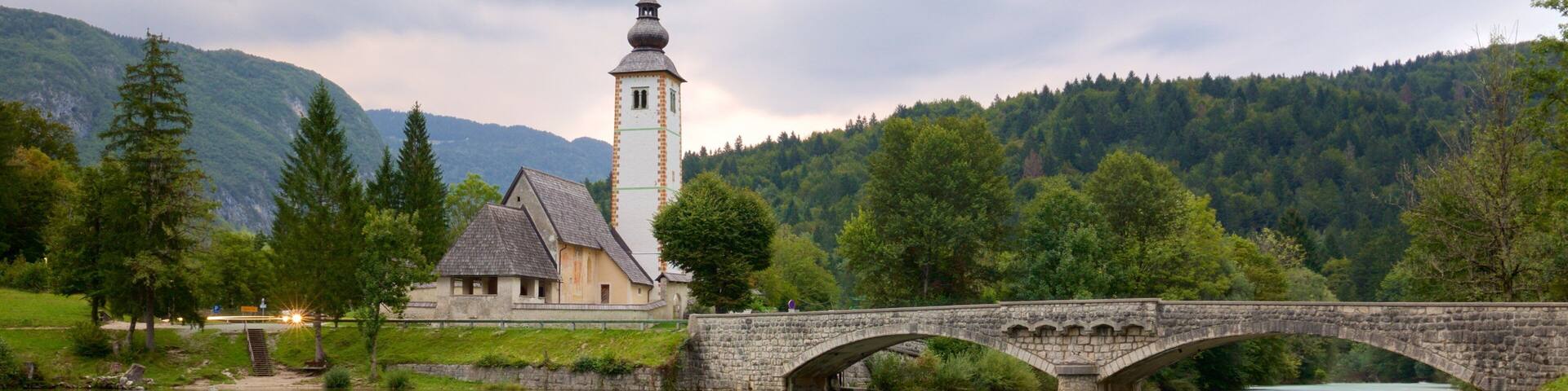 Un puente de piedra sobre un río tranquilo, una iglesia con un campanario y una ladera boscosa al fondo.