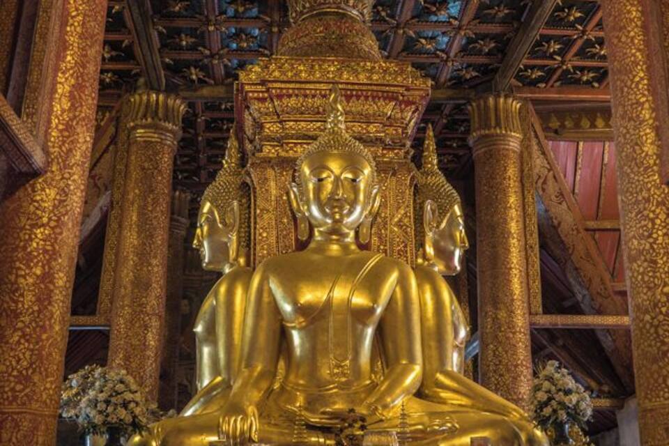 Una gran estatua dorada de Buda en un templo con elaboradas esculturas y columnas de madera.