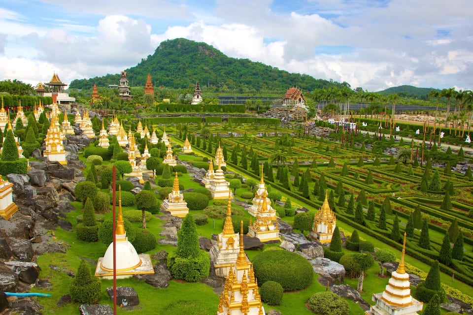 A temple complex with numerous golden spires and white structures, surrounded by lush greenery and a mountainous backdrop.