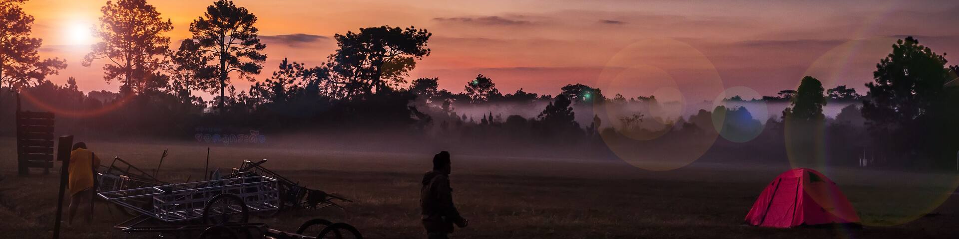 Un atardecer sobre un campo con una persona de pie cerca de un carrito y una tienda roja.