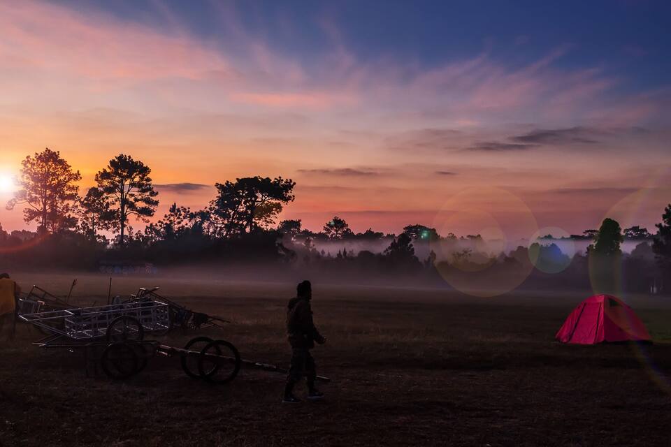 Un atardecer sobre un campo con una persona de pie cerca de un carrito y una tienda roja.