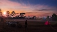 A sunset over a field with a person standing near a cart and a red tent.