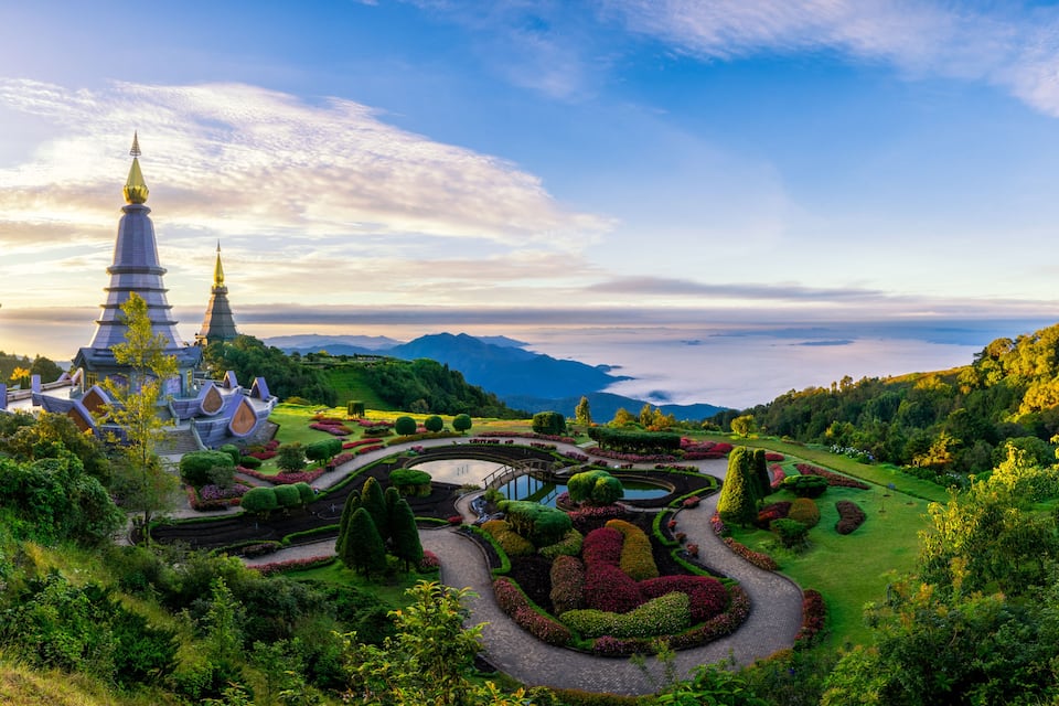 A temple complex with tiered spires, surrounded by lush gardens and a mountainous backdrop.