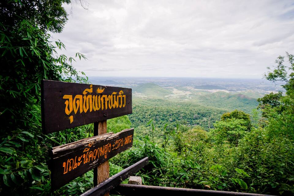 A wooden sign with Thai text and a view of a lush green valley.