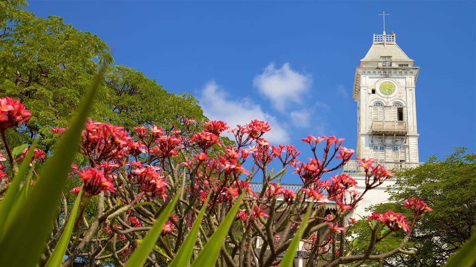 A clock tower with a cross on top, surrounded by trees and pink flowers.