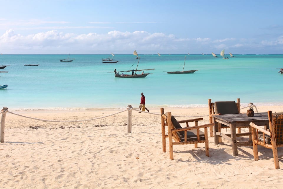 Ein Strand mit Holzbänken und -tischen, einer Seilsperre und mehreren Booten auf türkisfarbenem Wasser.