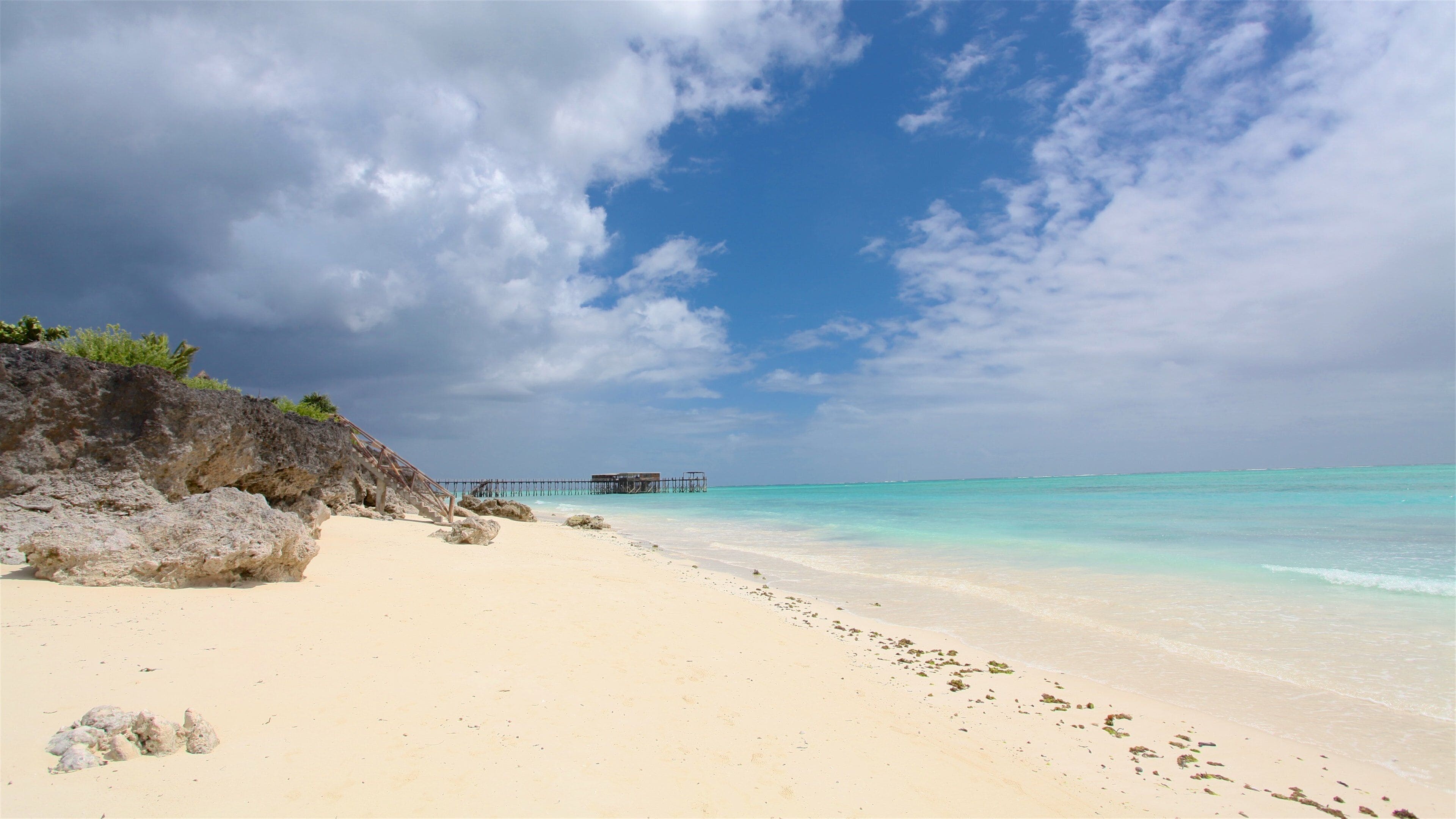 A beach with clear turquoise water, a rocky cliff, and a pier in the distance.