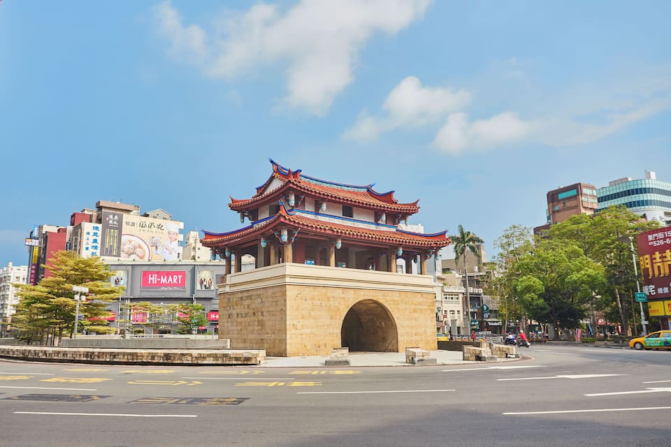 A traditional Chinese pavilion with a red roof and blue accents, surrounded by modern buildings and a clear blue sky.