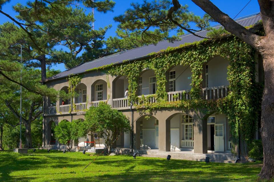 A historic building with arched windows and a balcony, surrounded by lush green trees and grass.