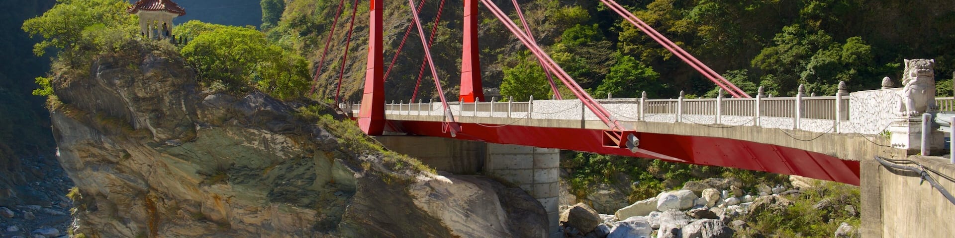 A red bridge spanning a rocky gorge with lush greenery and a temple on a cliff.
