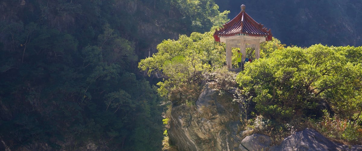 A traditional pavilion on a cliff edge with lush greenery and a mountainous backdrop.