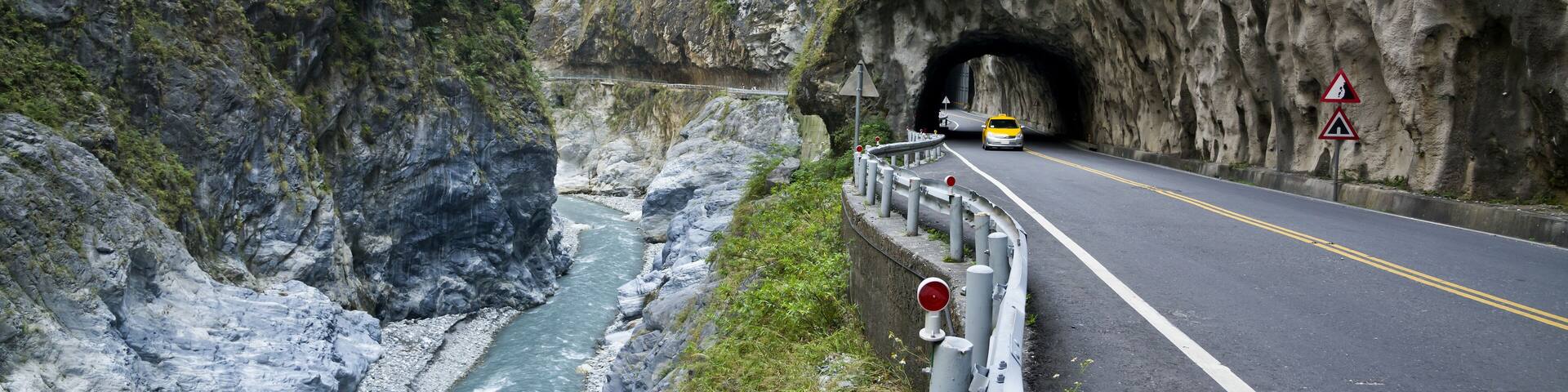 A road passing through a rocky canyon with a river on one side and a tunnel entrance on the other.