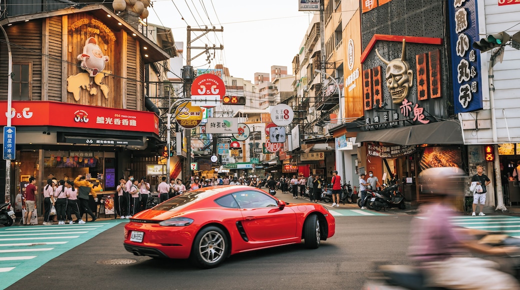 A busy urban street with a red sports car driving down the road, surrounded by various shops and signs.
