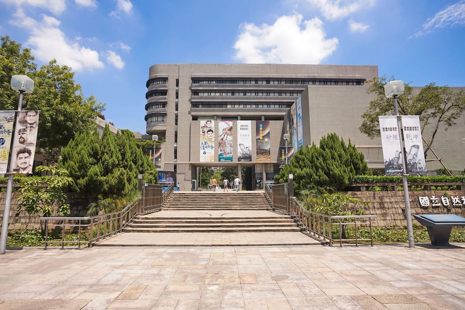A modern building with a staircase leading to the entrance, surrounded by greenery and banners.